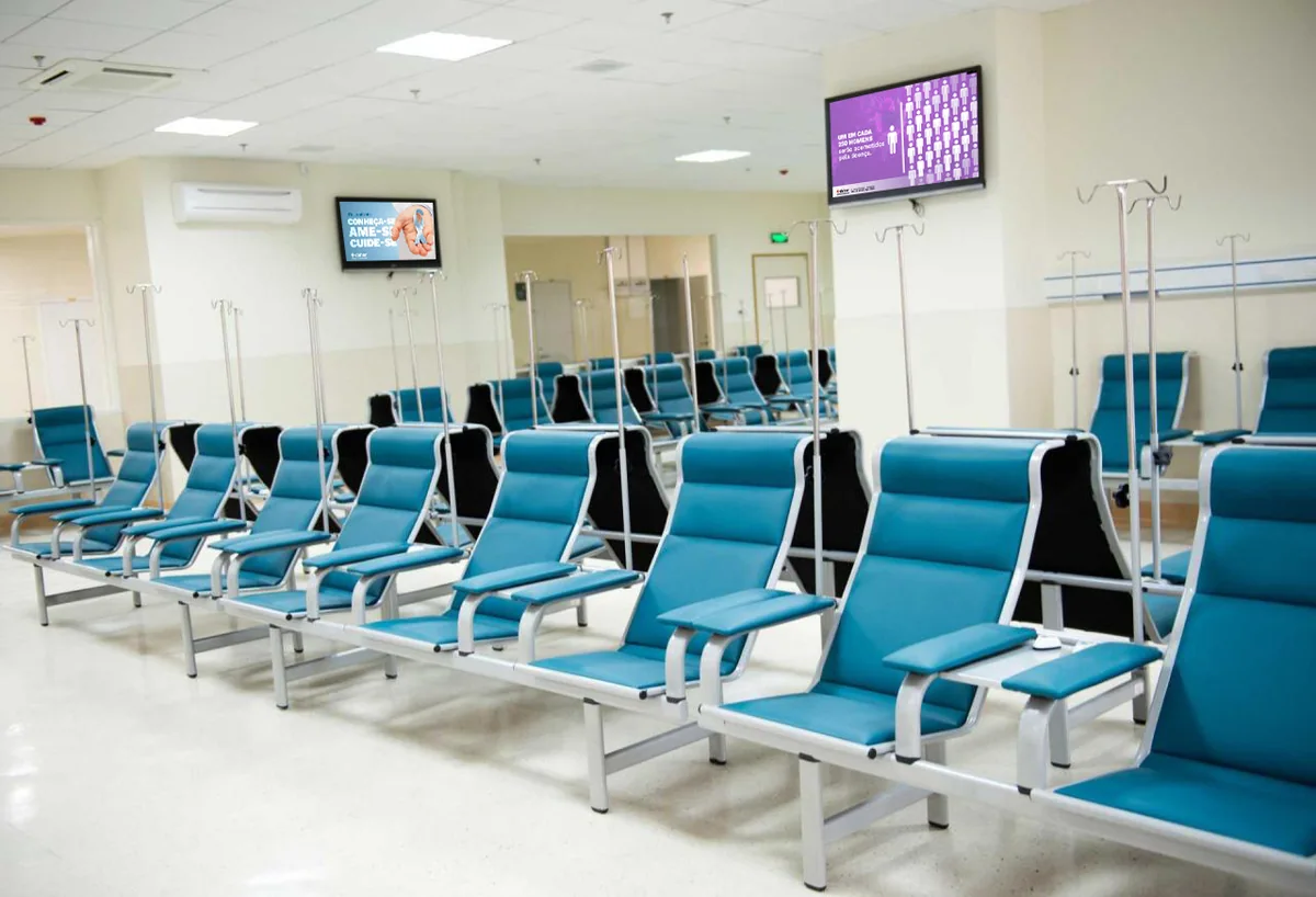 Sala de infusão do Daher Hospital Lago Sul com poltronas azuis e duas TVs exibindo as telas da campanha Novembro Azul