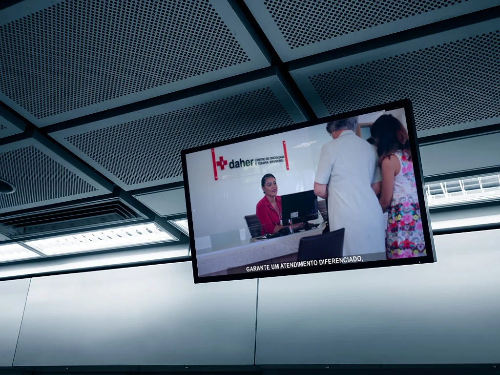 TV corporativa suspensa no teto da recepção do Centro de Oncologia e Terapia Infusional do Daher, com o vídeo institucional e a legenda garante um atendimento diferenciado