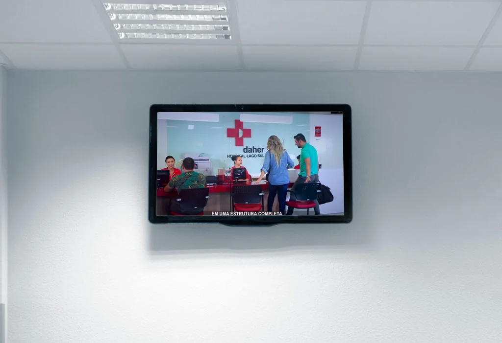 TV corporativa na parede da recepção do Daher Hospital Lago Sul, com o vídeo institucional em exibição e a legenda em uma estrutura completa